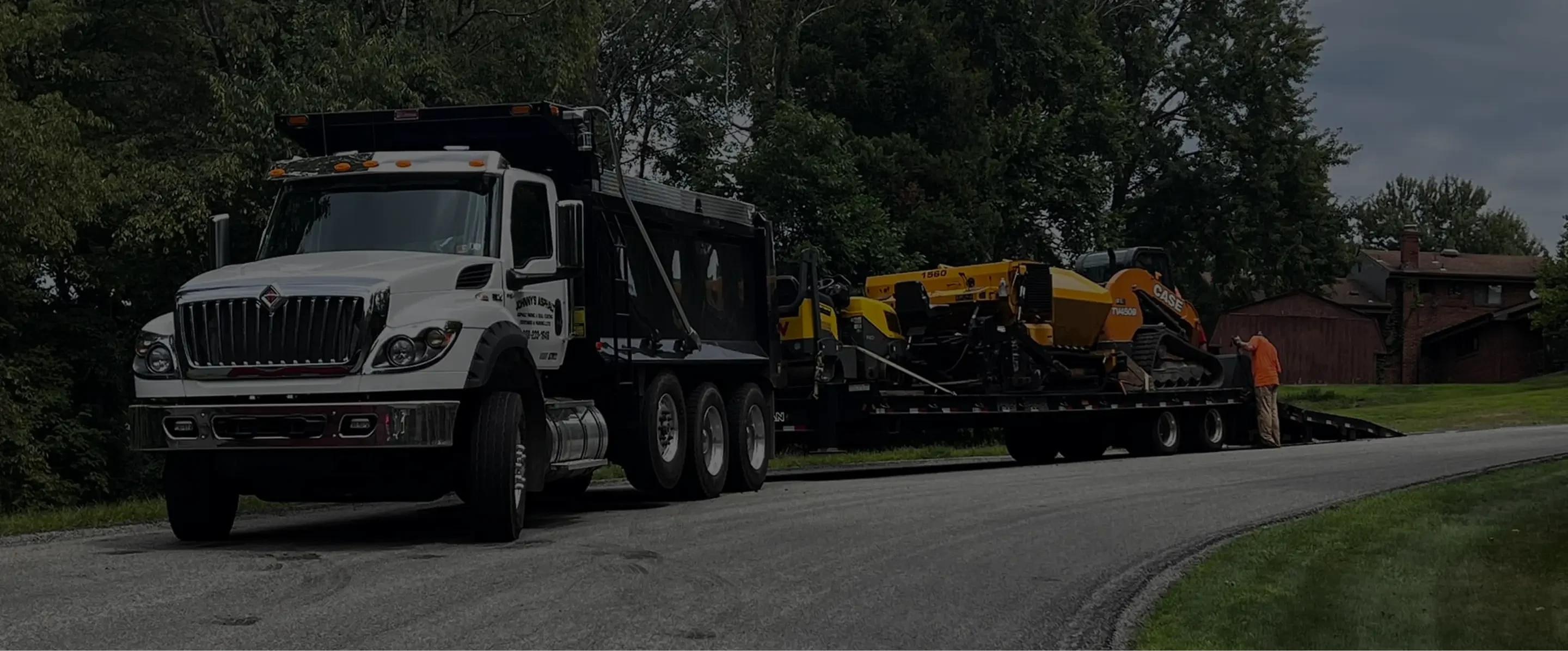 A white dump truck parked on a curved road, hauling construction machinery with a worker adjusting equipment on the trailer.