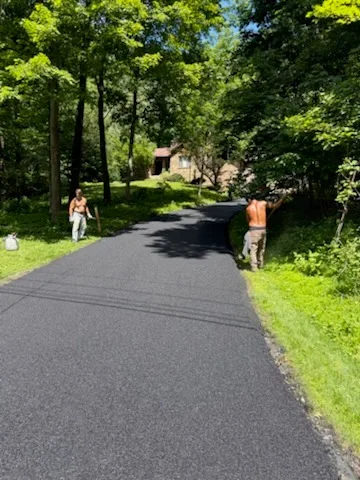 Newly paved black asphalt driveway lined with mature trees and green lawn